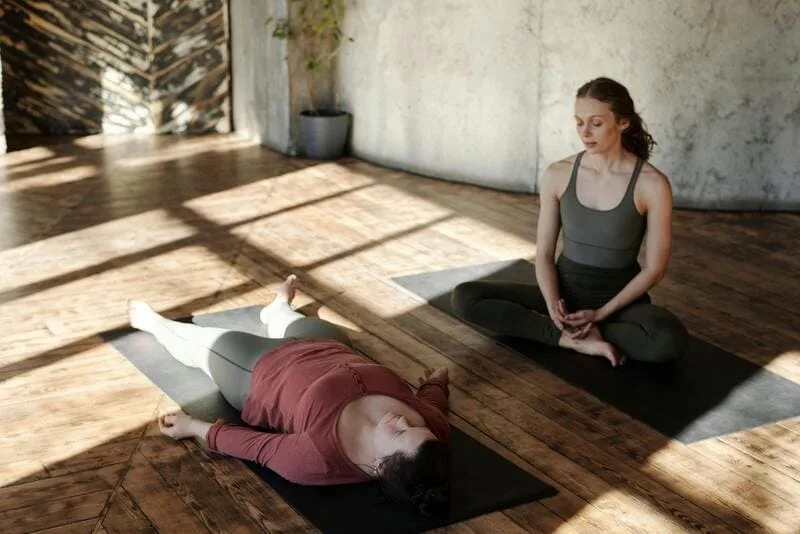 A woman lying on a mat with another woman guiding her meditation