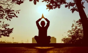 Silhouette of a person meditating at sunrise, hands in prayer position above their head, with the sun shining in the background
